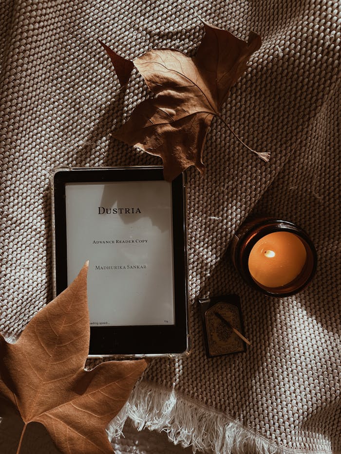 Warm autumn scene with tablet, candle, and dried leaves on a textured blanket.