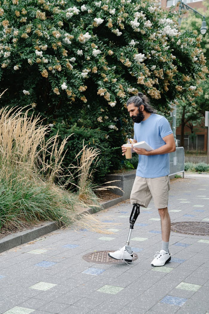 A man with a prosthetic leg walks outdoors holding a cup and notebook, surrounded by greenery.